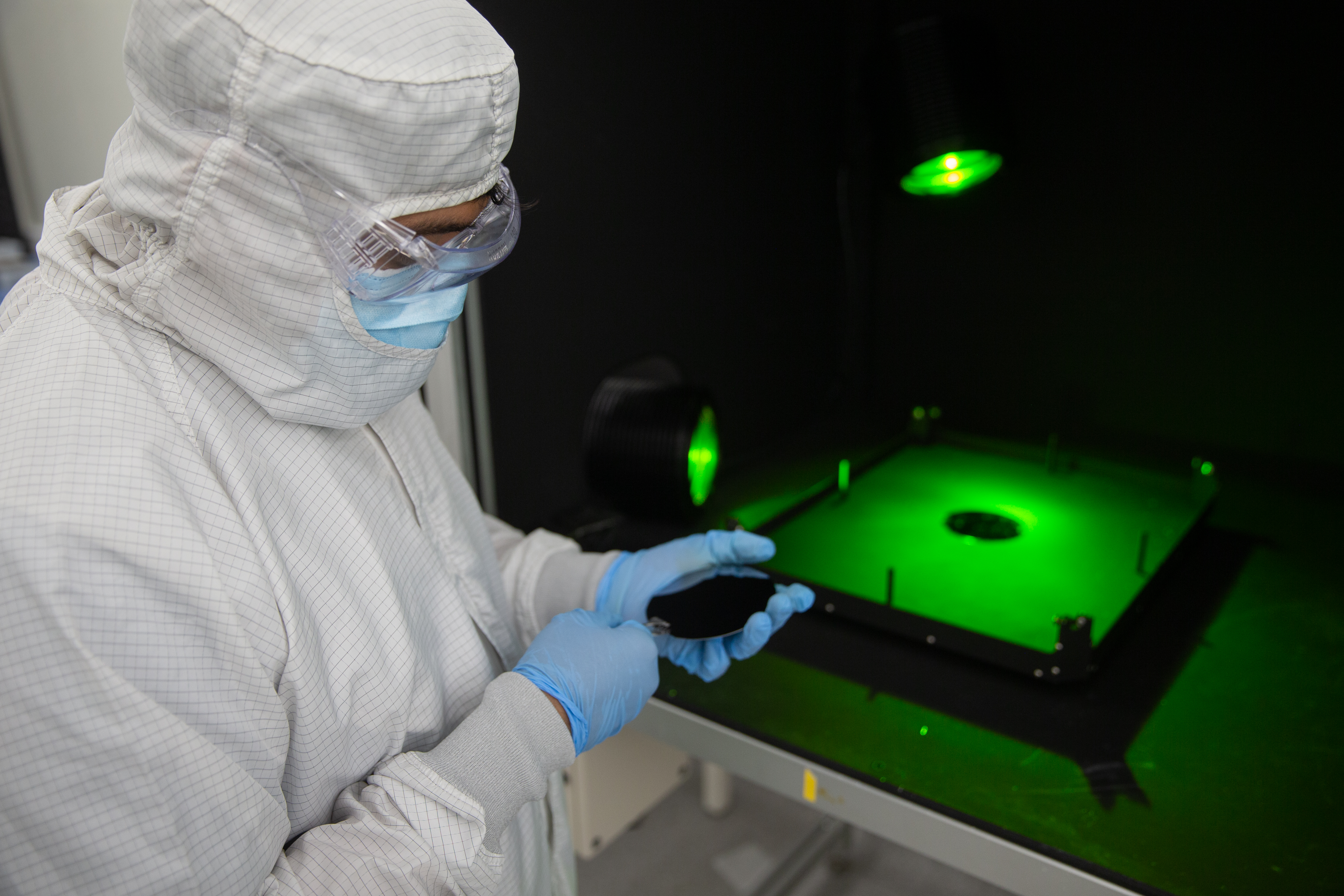 Cleanroom technician using precision tools to handle a semiconductor wafer under green inspection lighting