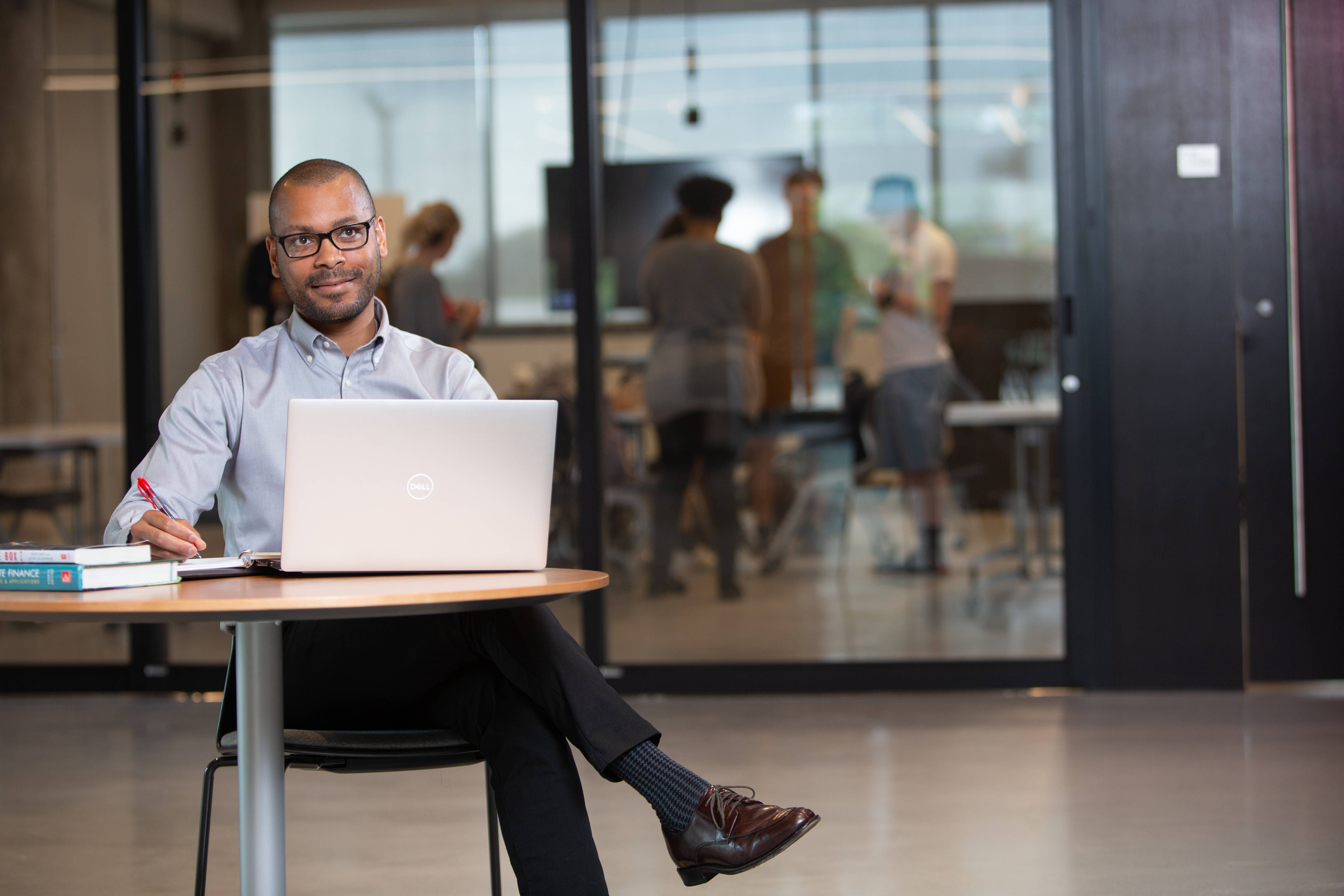 A professional working on a laptop at a table with a team meeting in the background.