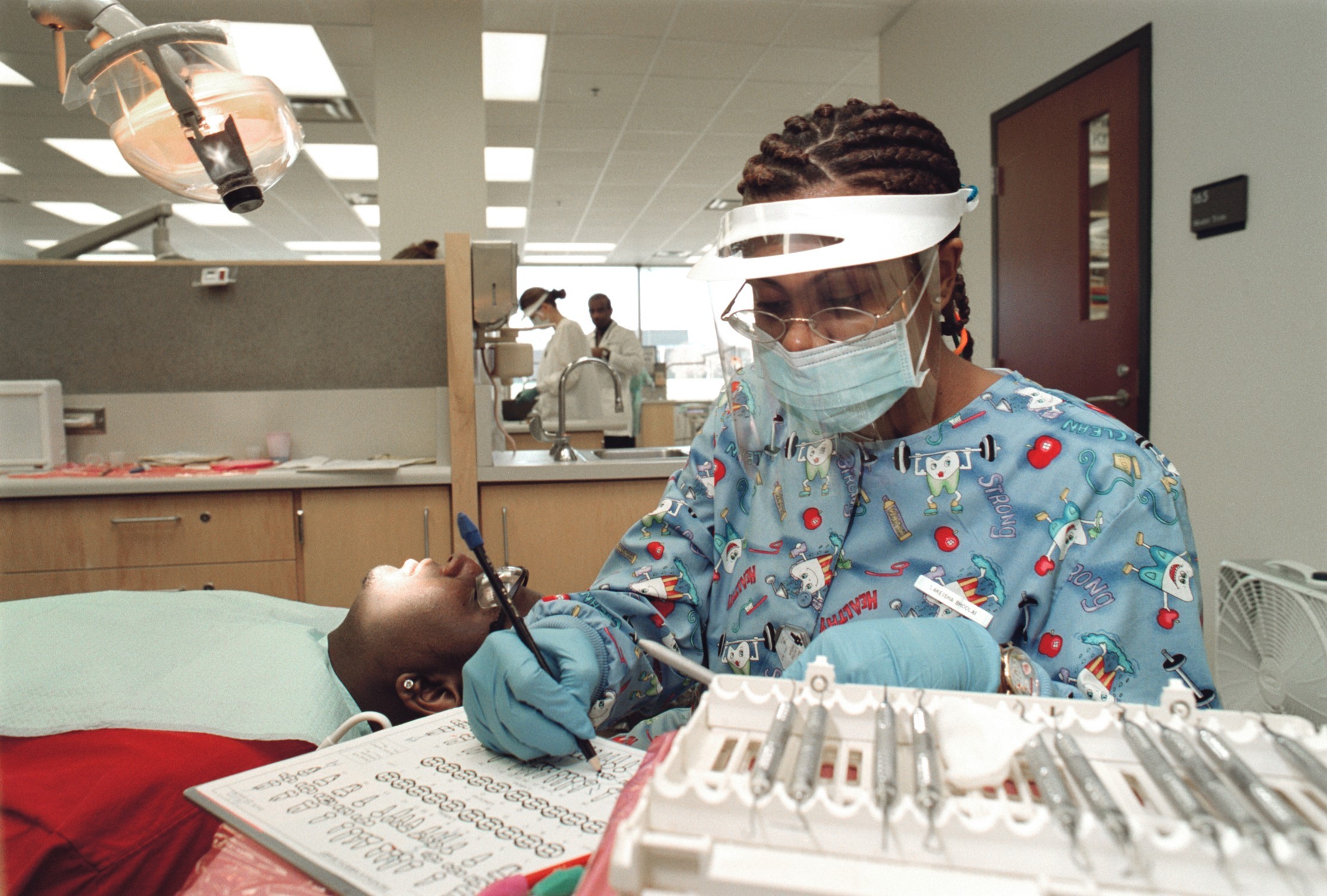 Dental professional wearing protective equipment documenting a patient’s oral exam while the patient receives care in a clinical setting