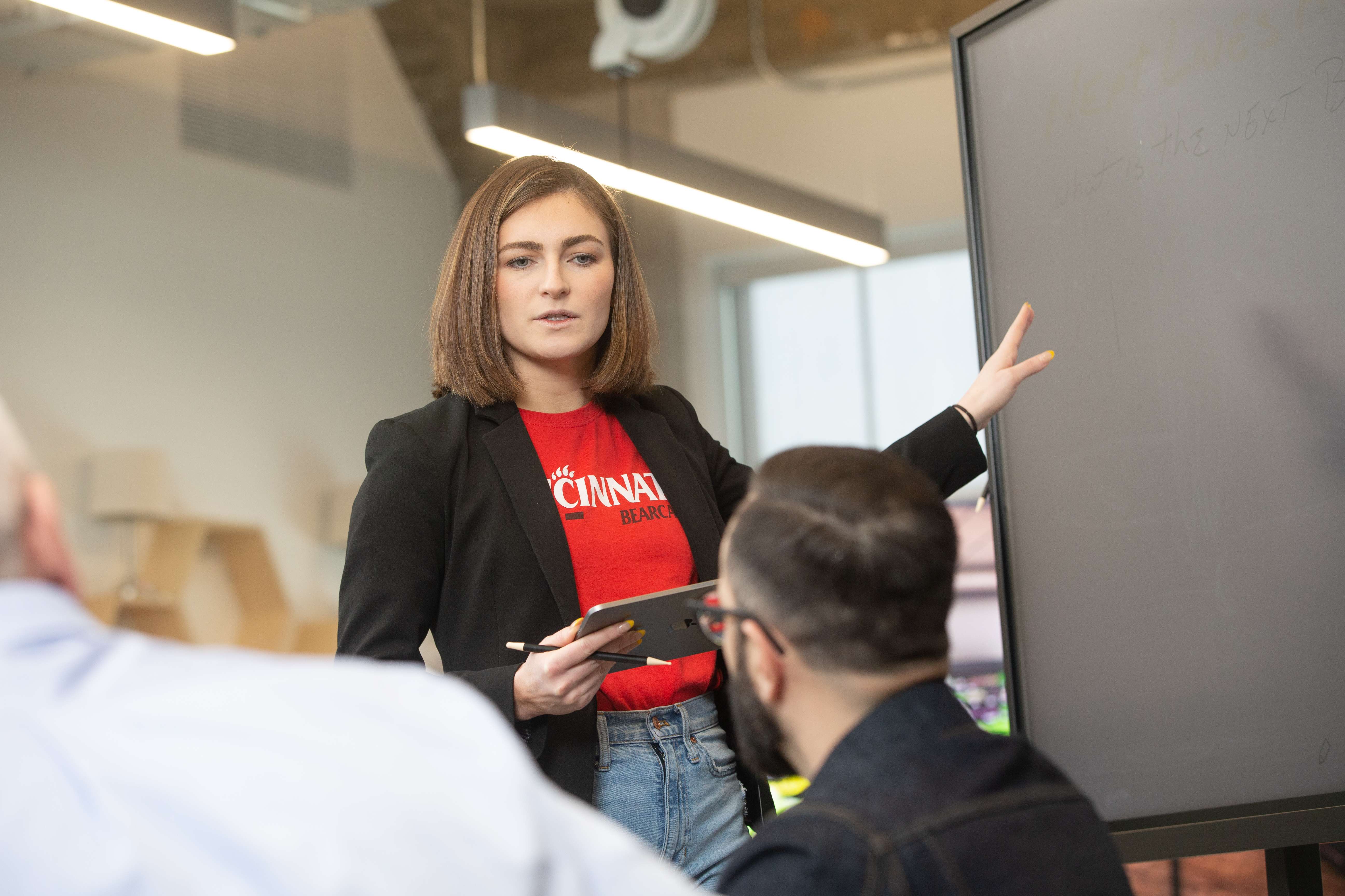 Person presenting to a group while pointing at a screen and holding a tablet