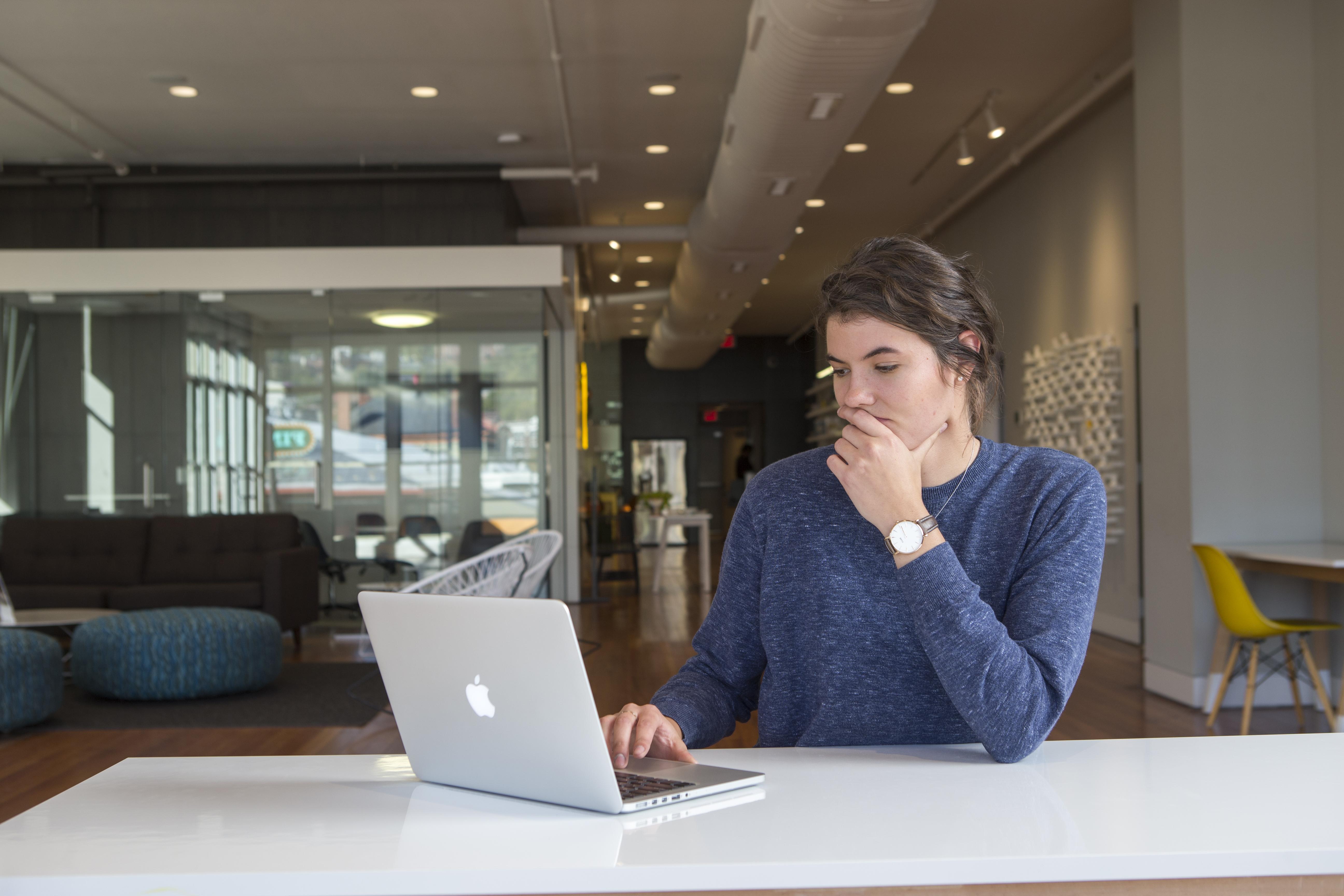 Person thoughtfully working on a laptop at a table in a modern, open workspace