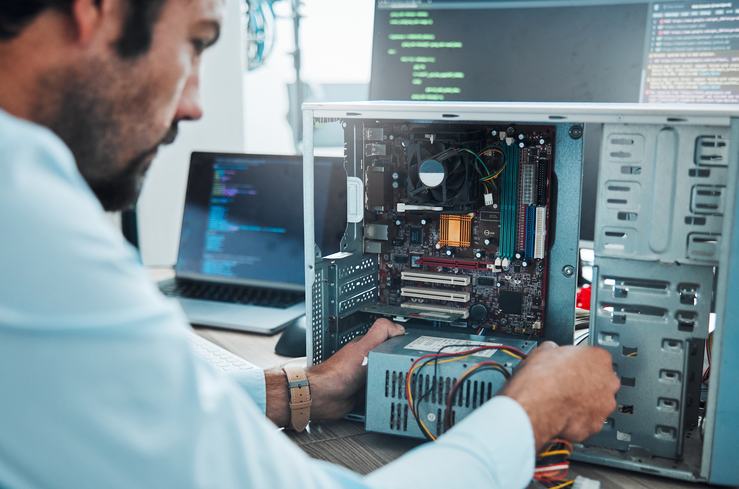 A learner installs or troubleshoots hardware inside an open desktop computer while working at a workstation with computer code displayed on nearby screens
