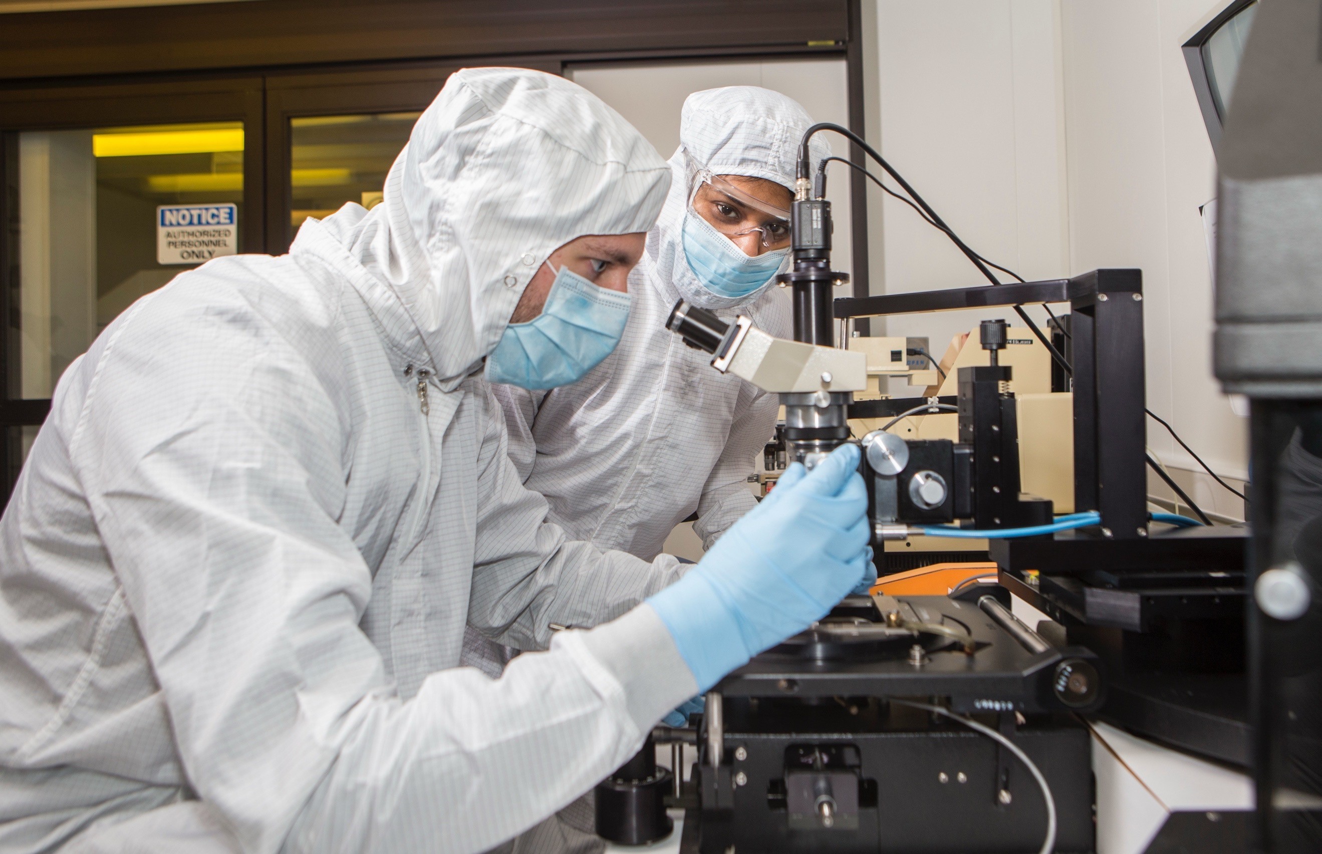Two technicians in cleanroom attire using a microscope to inspect semiconductor equipment