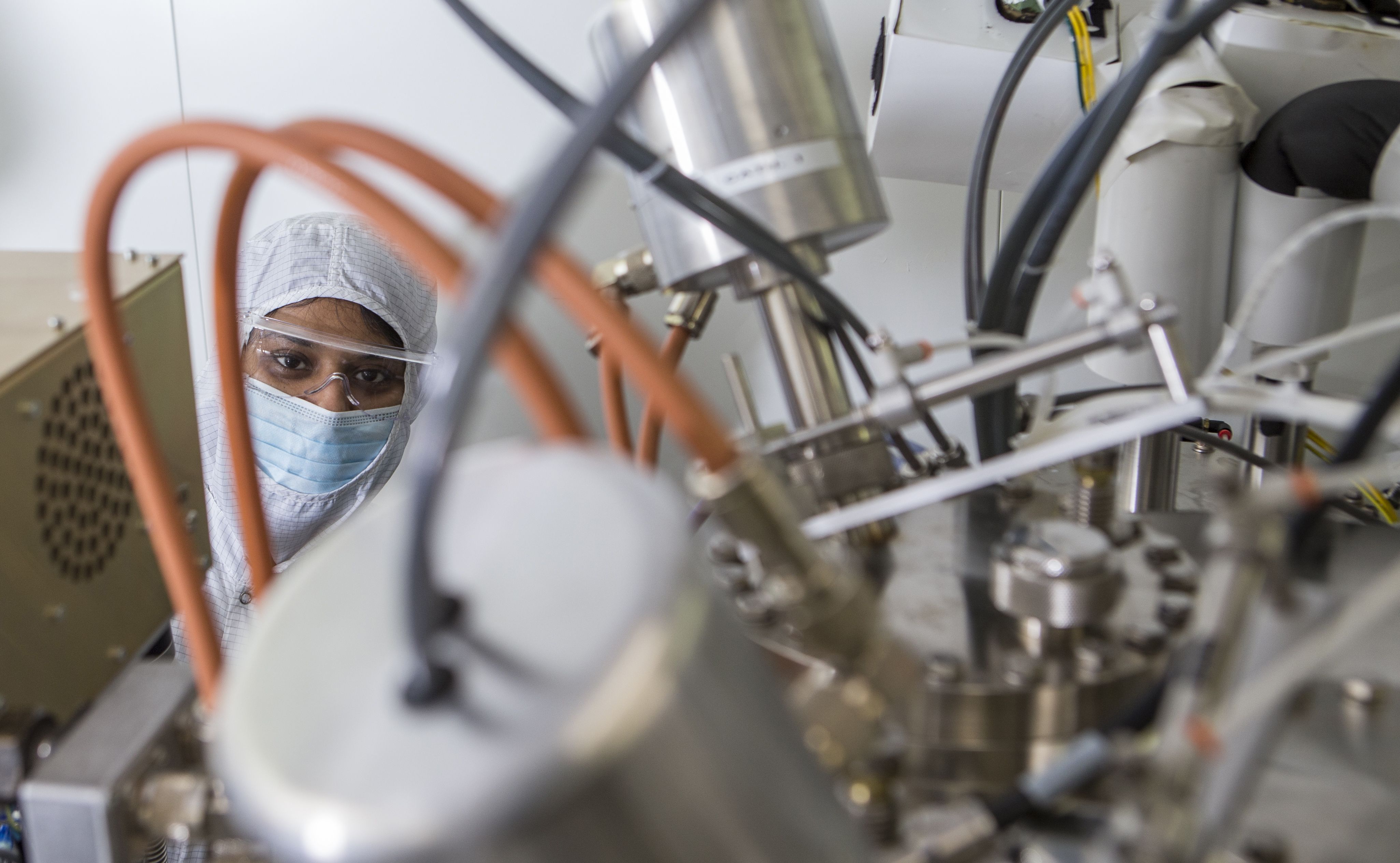 A technician wearing cleanroom protective equipment inspects semiconductor vacuum and gas handling equipment in a semiconductor manufacturing cleanroom